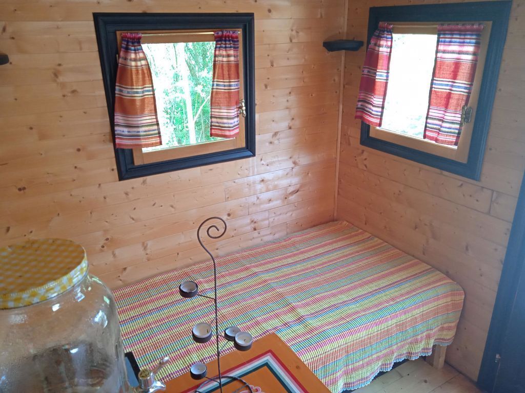 Una habitación de cabaña pequeña y de madera con dos ventanas, cada una con una cortina a rayas. Hay una cama con una colcha de rayas coloridas y una mesita con un tarro y velas.