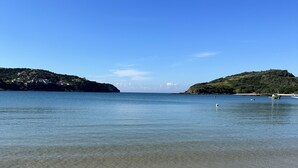 Une plage avec une eau claire et peu profonde, une vue lointaine sur des collines et quelques bateaux.