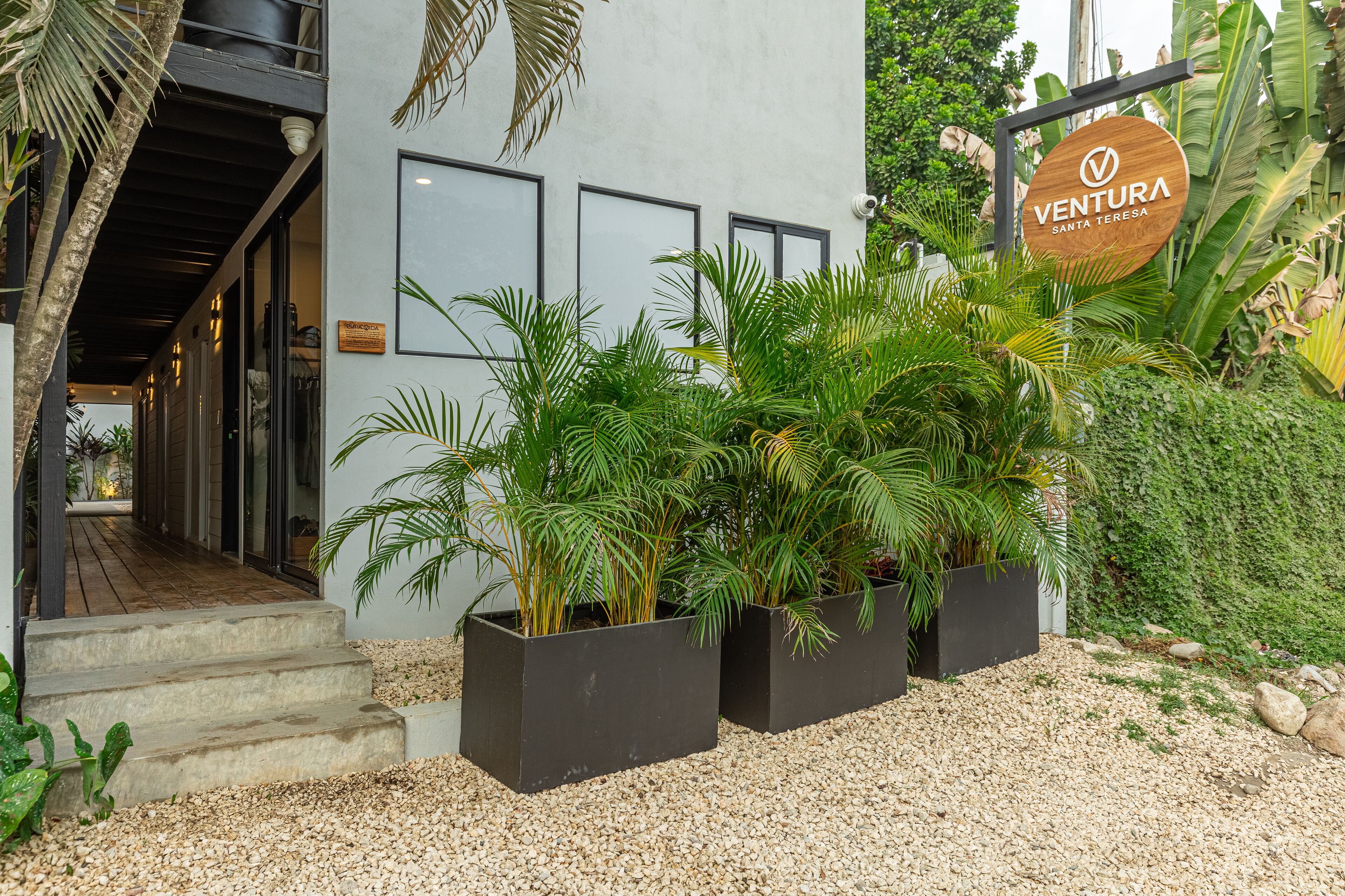 Entrada de un edificio con un letrero que dice "VENTURA SANTA TERESA", rodeada de vegetación exuberante y un camino de grava.