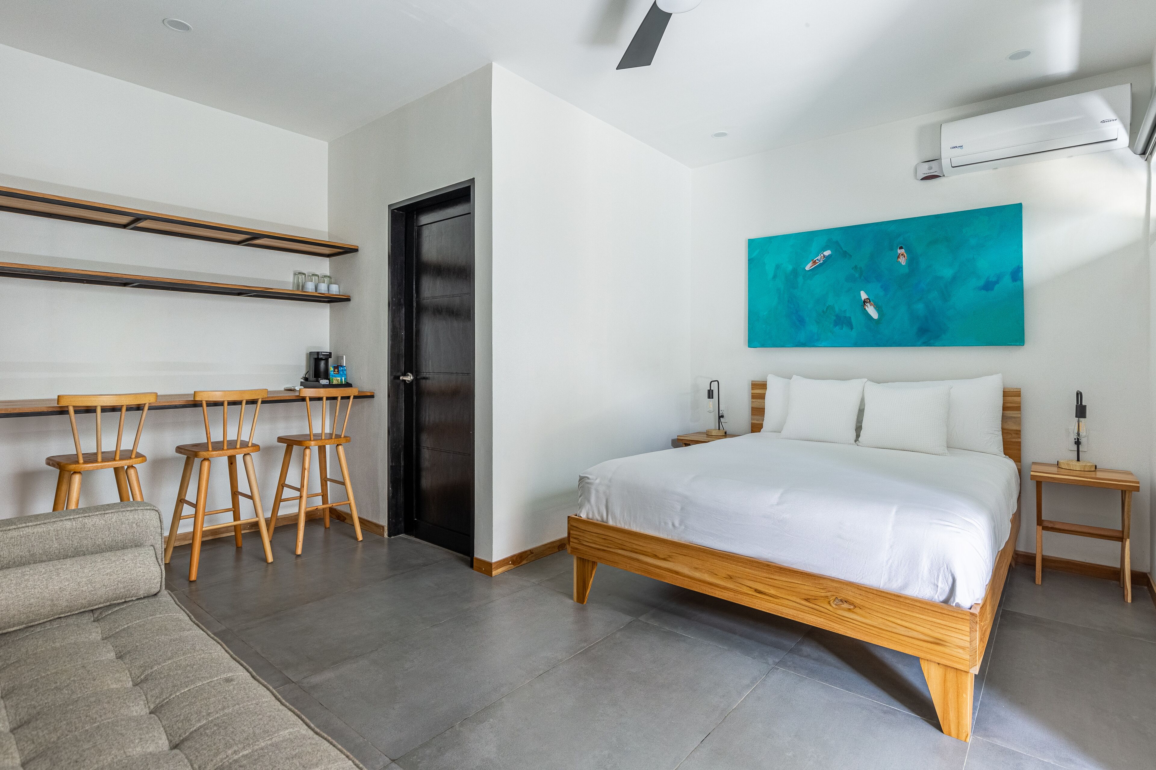 A modern bedroom with a large bed, a desk with chairs, a coffee machine, and a painting on the wall.
