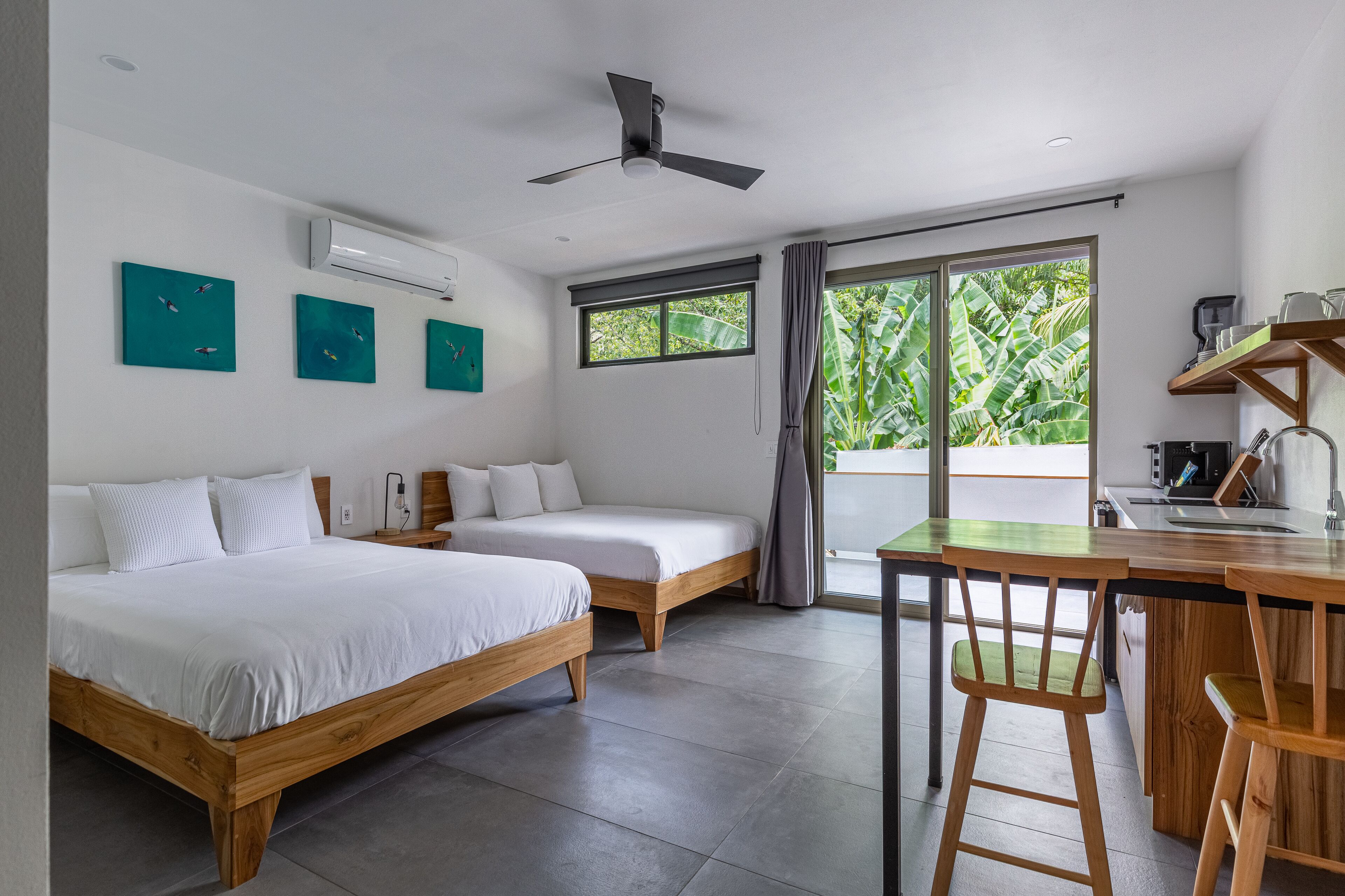 Una habitación de hotel moderna con dos camas individuales, un escritorio de madera con una silla, un ventilador de techo y un ventanal con vistas a zonas verdes.