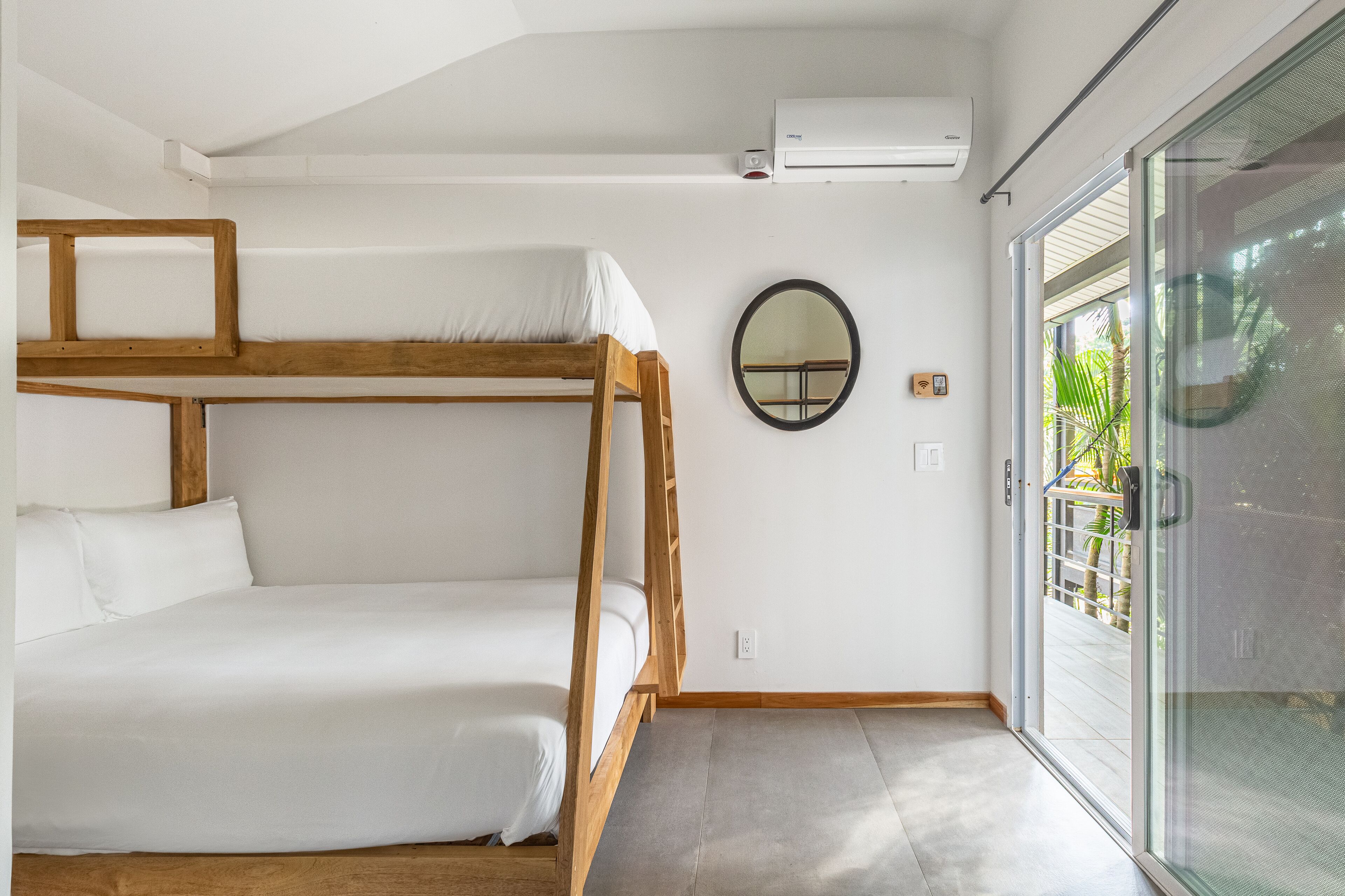 A bunk bed room with a wooden frame, white bedding, a round mirror, and a sliding glass door leading to a balcony with plants.
