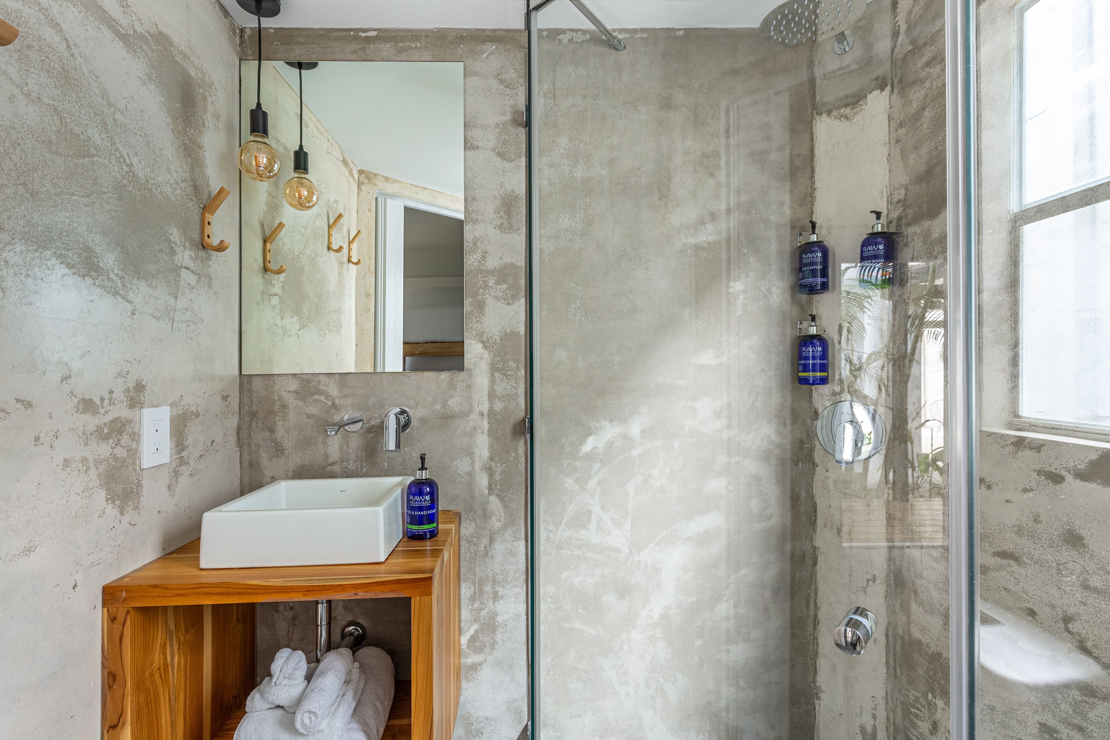 A bathroom with a concrete wall, a wooden vanity, a white sink, and a glass shower enclosure.