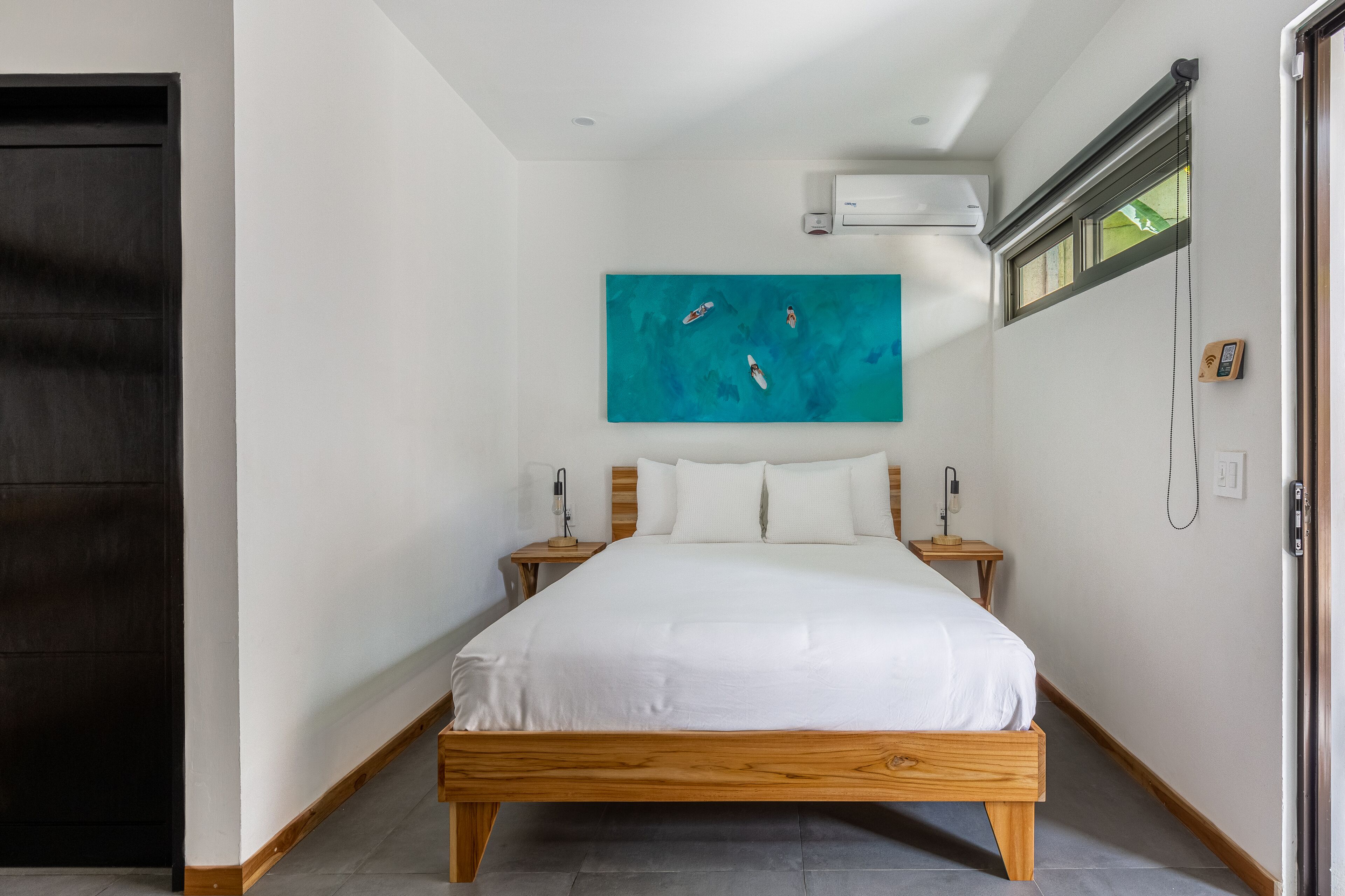 A bedroom with a wooden bed, white bedding, a teal artwork above the bed, a nightstand with a lamp, and a window with blinds.