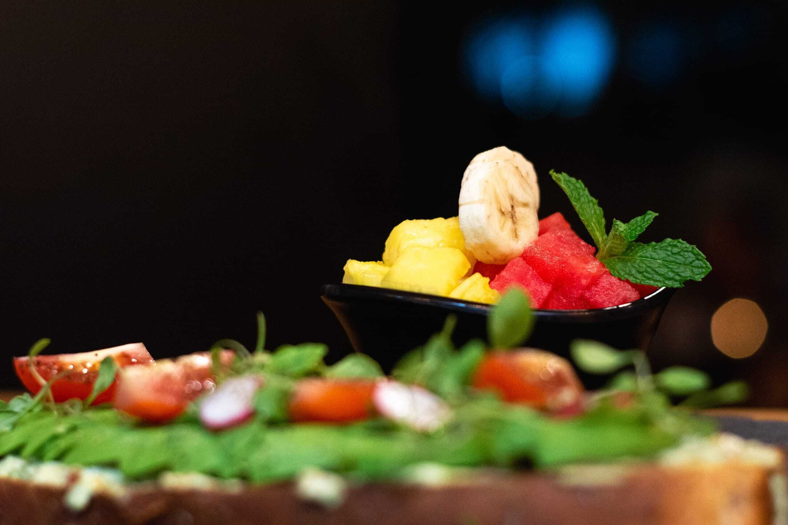 A bowl of fruit salad with bananas, watermelon, and pineapple on a bed of greens, with a mint leaf garnish.