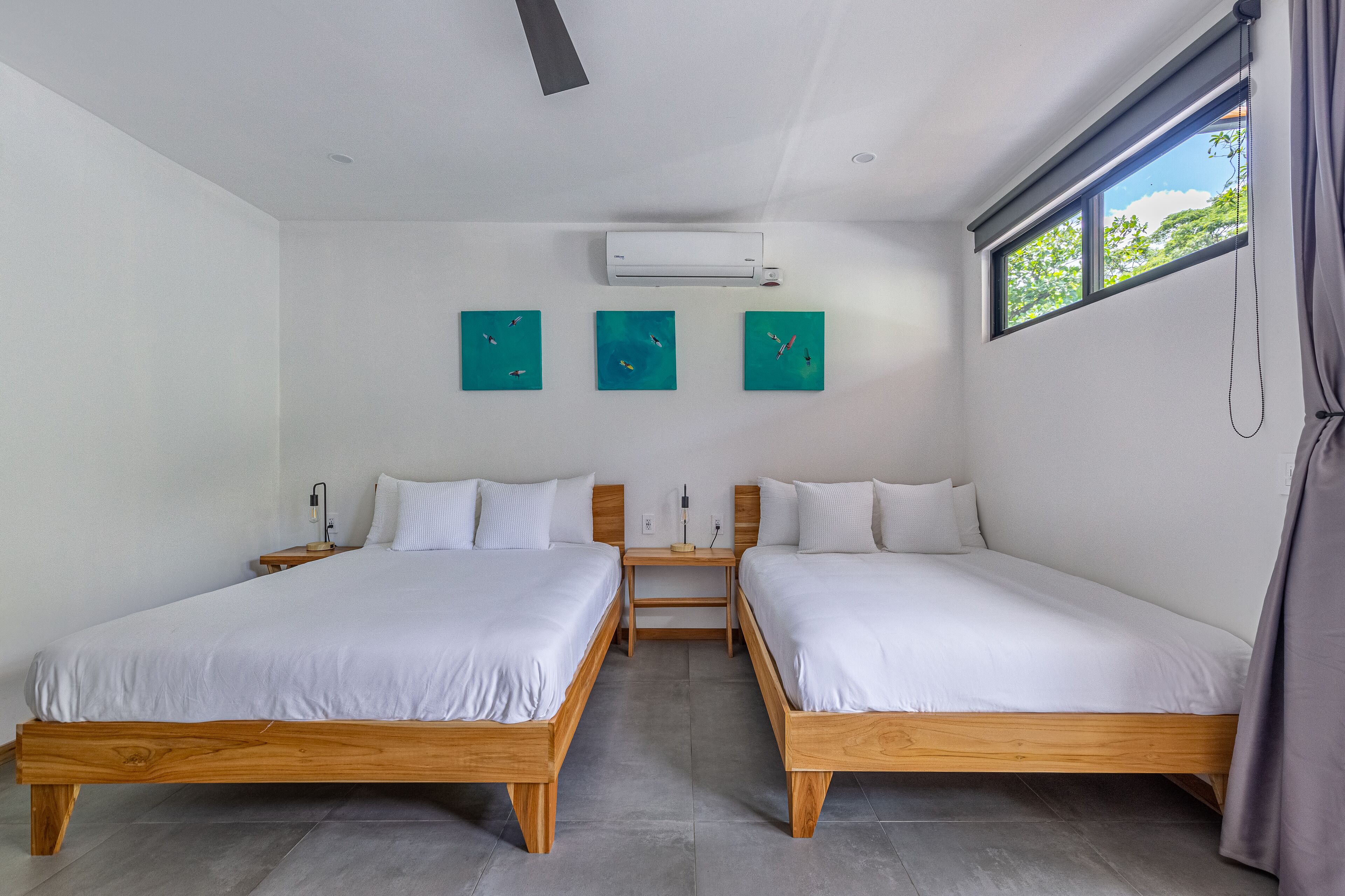 A modern bedroom with two wooden beds, a ceiling fan, an air conditioner, and three teal artwork pieces on the wall.