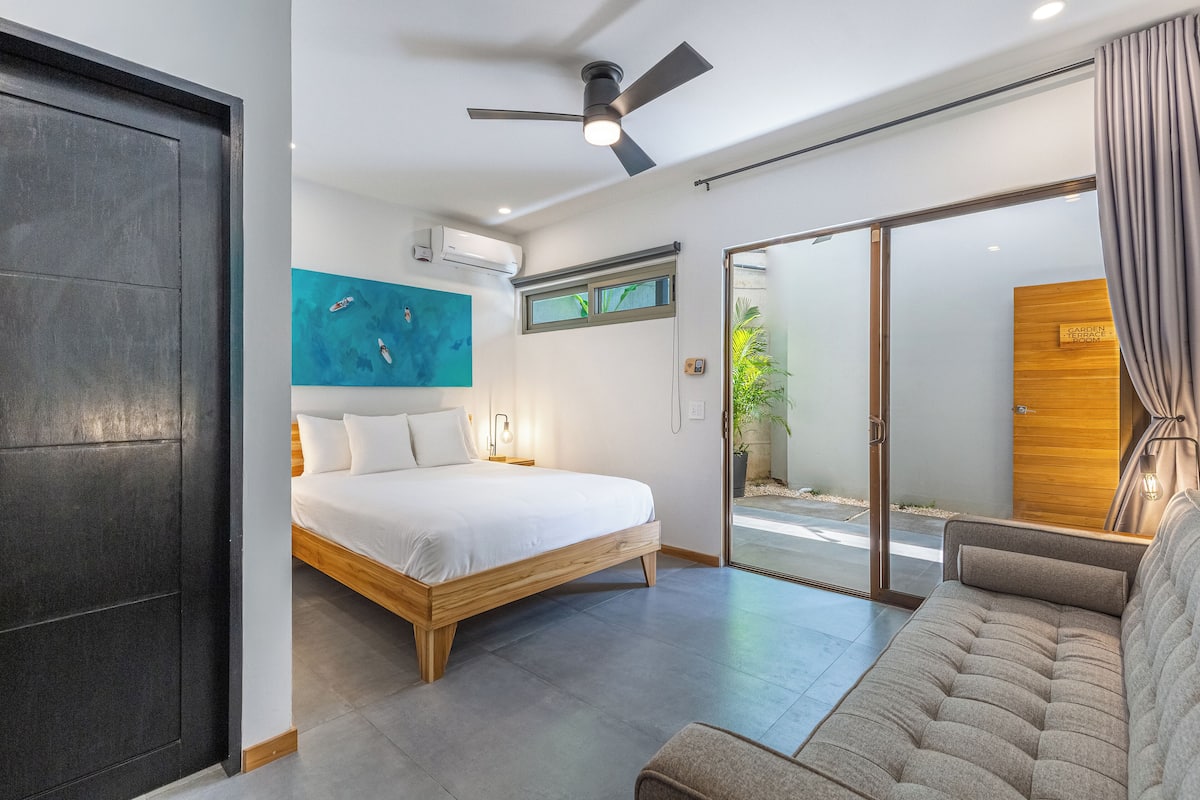 Un dormitorio moderno con cama, sofá, ventilador de techo y puerta corredera de cristal que da a un espacio exterior.