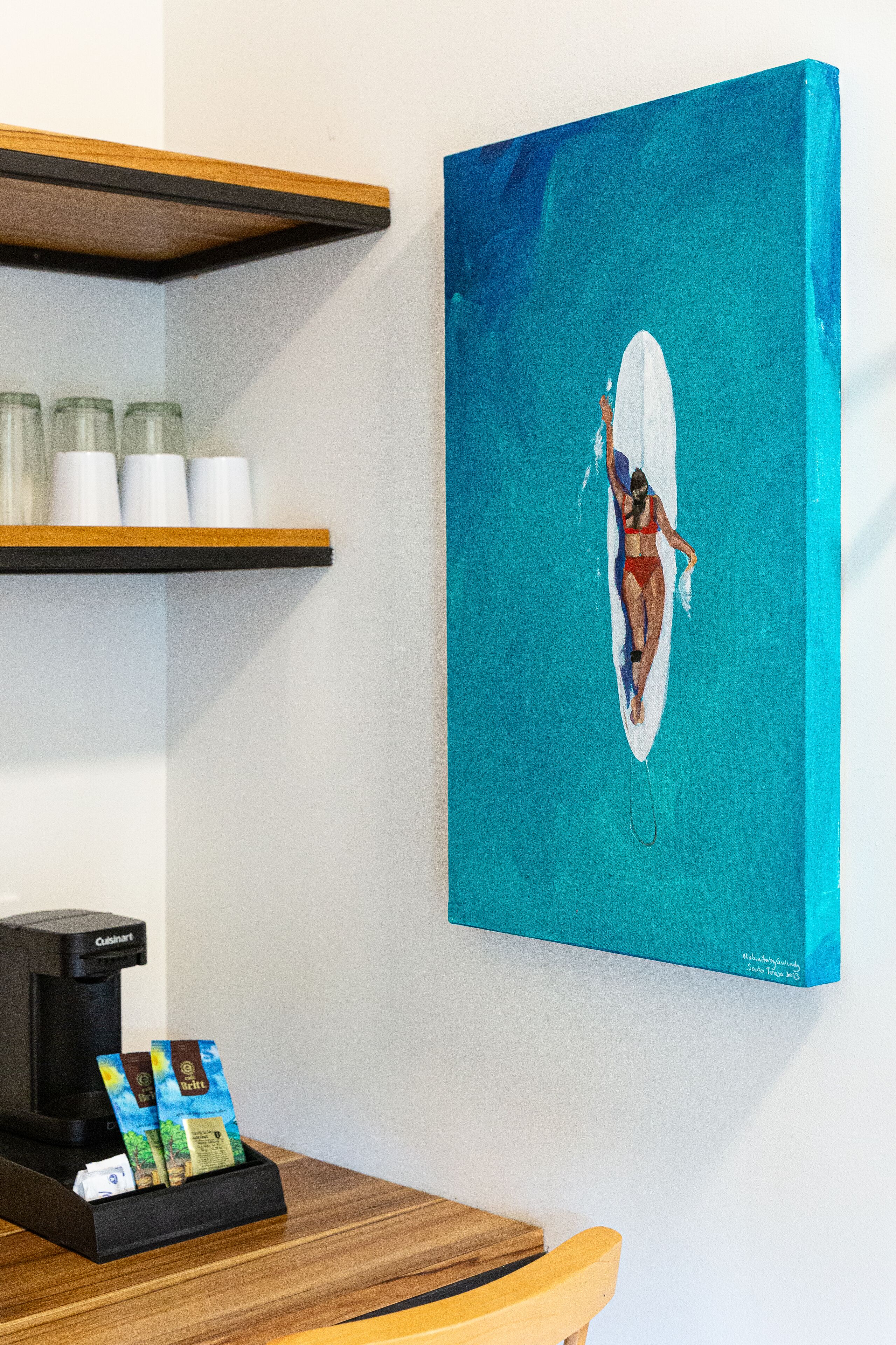 Un tableau représentant une femme sur une planche de surf dans une piscine, une machine à café et une étagère avec des bocaux.