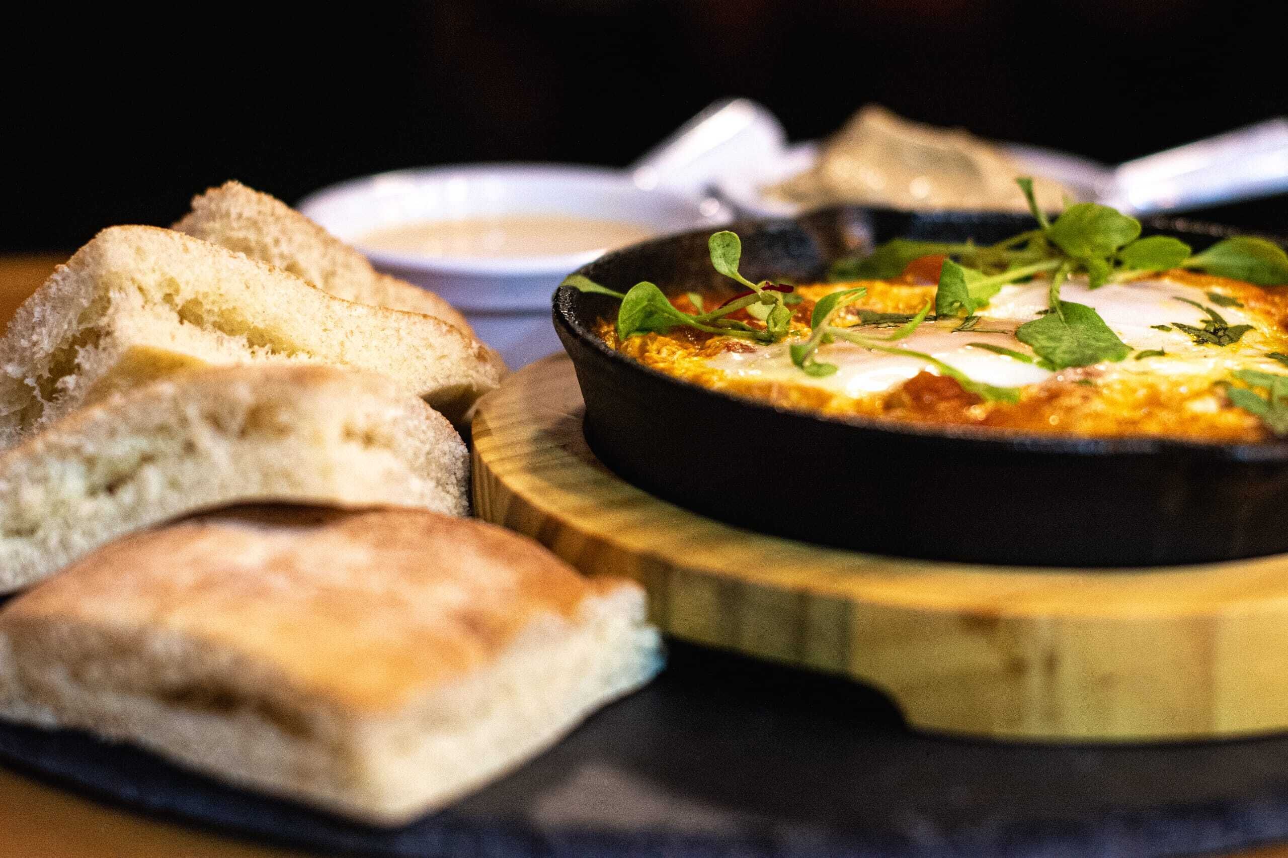 A dish of baked eggs with greens, accompanied by slices of bread on a wooden board.