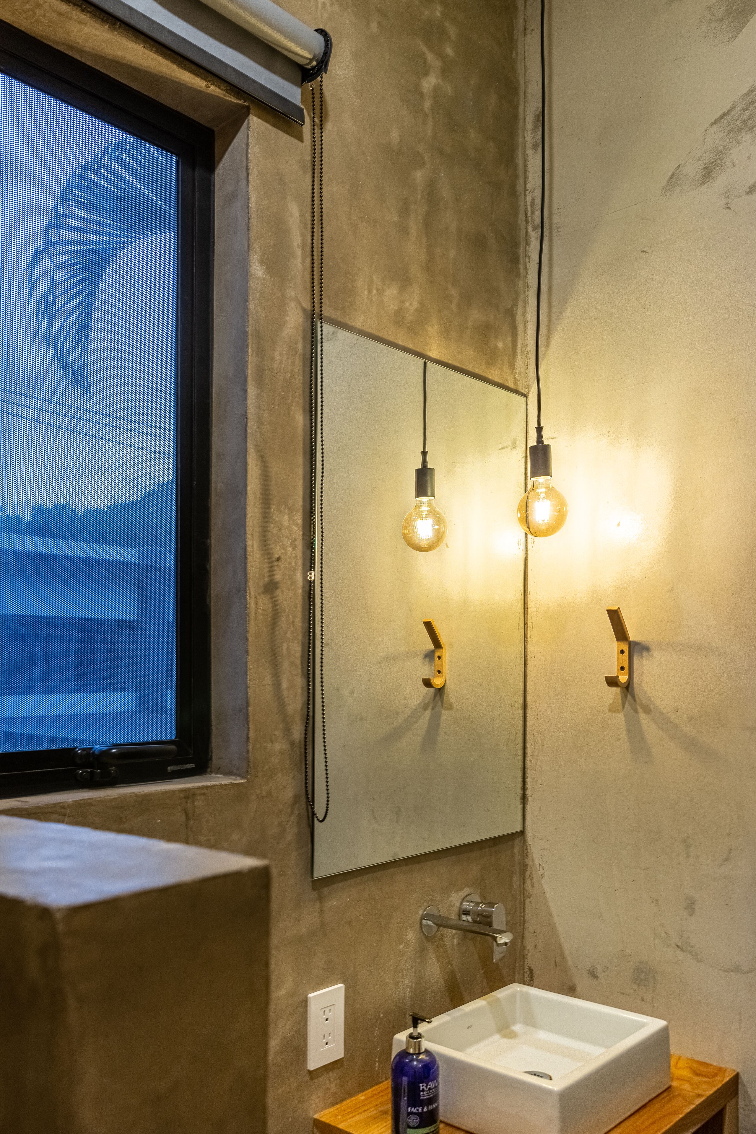 A bathroom with a large mirror, two hanging light bulbs, a white sink, and a window with a view of mountains.
