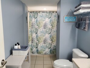 Casa de banho com cortina de chuveiro estampada com padrão de folhagem tropical, sanita, lavatório e porta-toalhas com toalhas.
