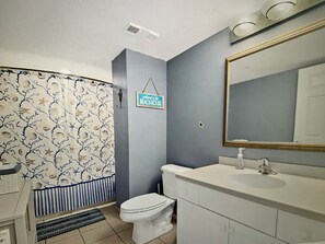 Une salle de bain avec un rideau de douche orné d’un motif de plage, des toilettes, un lavabo avec un miroir et une pancarte sur laquelle on peut lire : « Bienvenue à la maison de plage ».