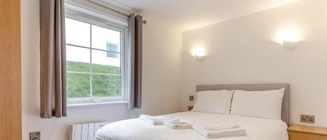 Habitación de hotel con cama, ventana con cortinas, radiador y lámpara fijada a la pared.