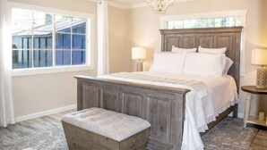 A bedroom with a large bed, a wooden bench, two bedside tables with lamps, and a window with a view of a blue building.