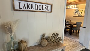 A wooden bench with a sign reading "LAKE HOUSE" above it, a vase with pampas grass, and a 'hello' sign.