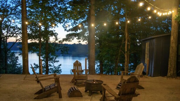 A rustic outdoor seating area with Adirondack chairs, a fire pit, and string lights, overlooking a lake.
