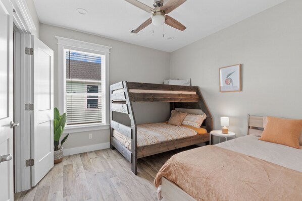 A bedroom with a bunk bed, a single bed, a ceiling fan, a bedside table with a lamp, a window with blinds, and a framed picture on the wall. - BRAND NEW! Sea Isles Cove - Steps From The Beach (Galveston)