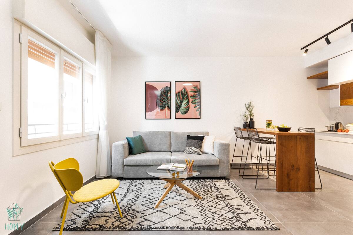 Un salon moderne avec un canapé gris, un fauteuil jaune et une table basse. Deux œuvres d’art représentant des feuilles sont encadrées et accrochées au mur. On y trouve également un coin repas avec une table et des chaises en bois, ainsi qu’un coin cuisine avec un comptoir et des placards.