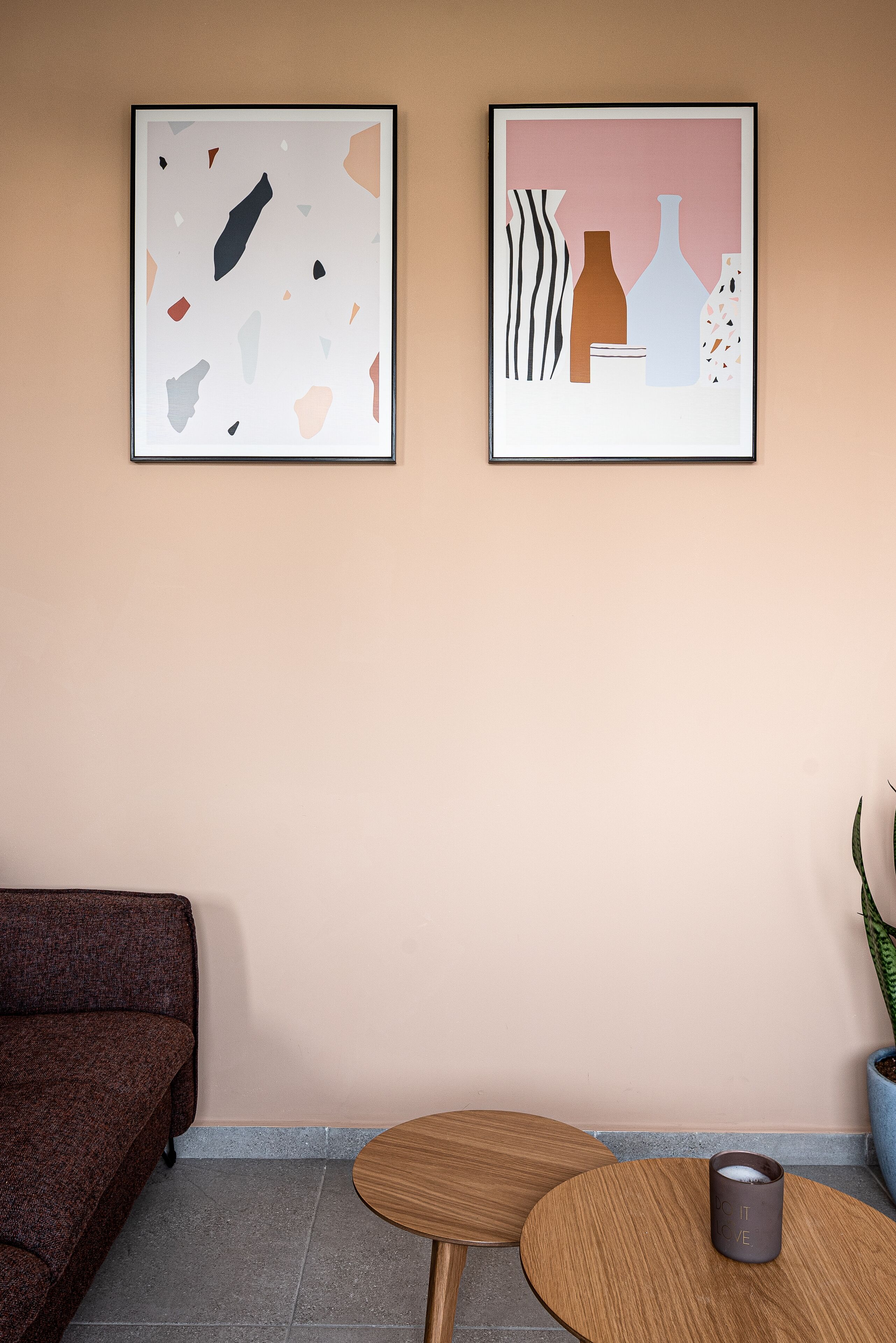 Una habitación con un sofá marrón, dos cuadros abstractos enmarcados, una mesita de madera con una taza y una planta en maceta.