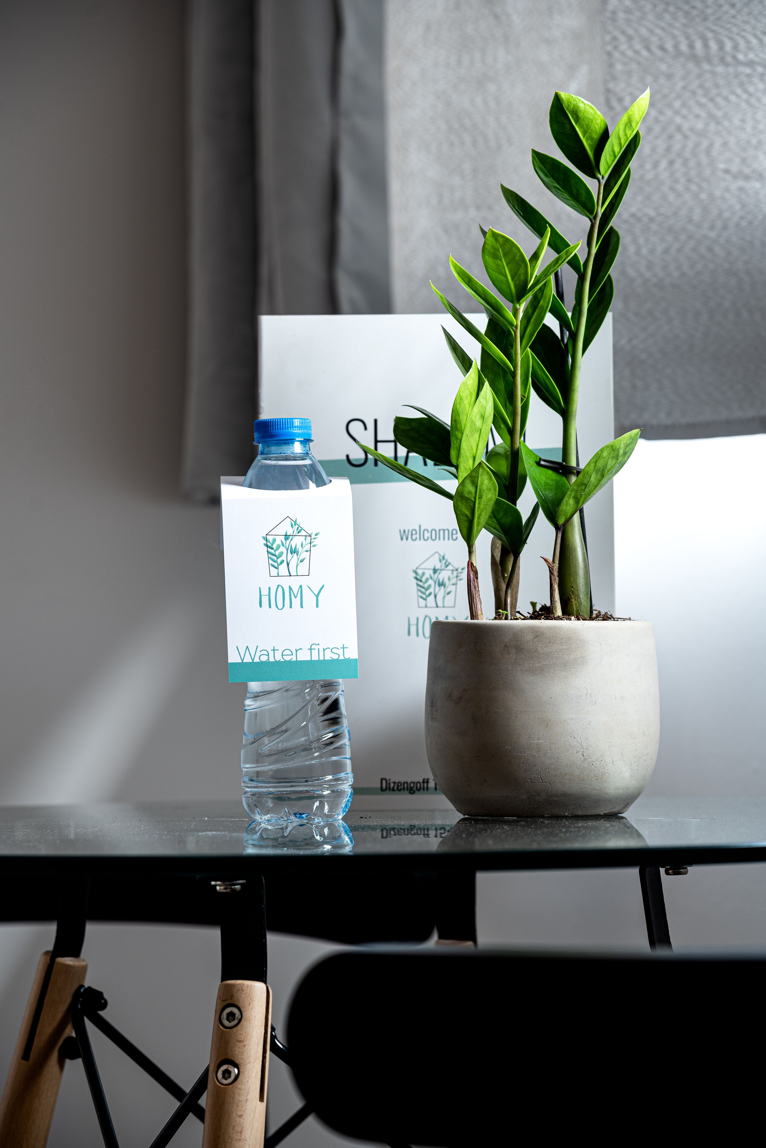 Une bouteille d’eau avec une étiquette, une plante en pot et un panneau portant le mot « bienvenue » sur une table.