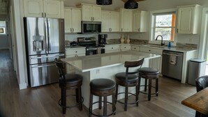 A modern kitchen with white cabinets, stainless steel appliances, a central island with bar stools, and a wooden ceiling. - Yellowstone Park Riverfront Chalet (Gardiner)