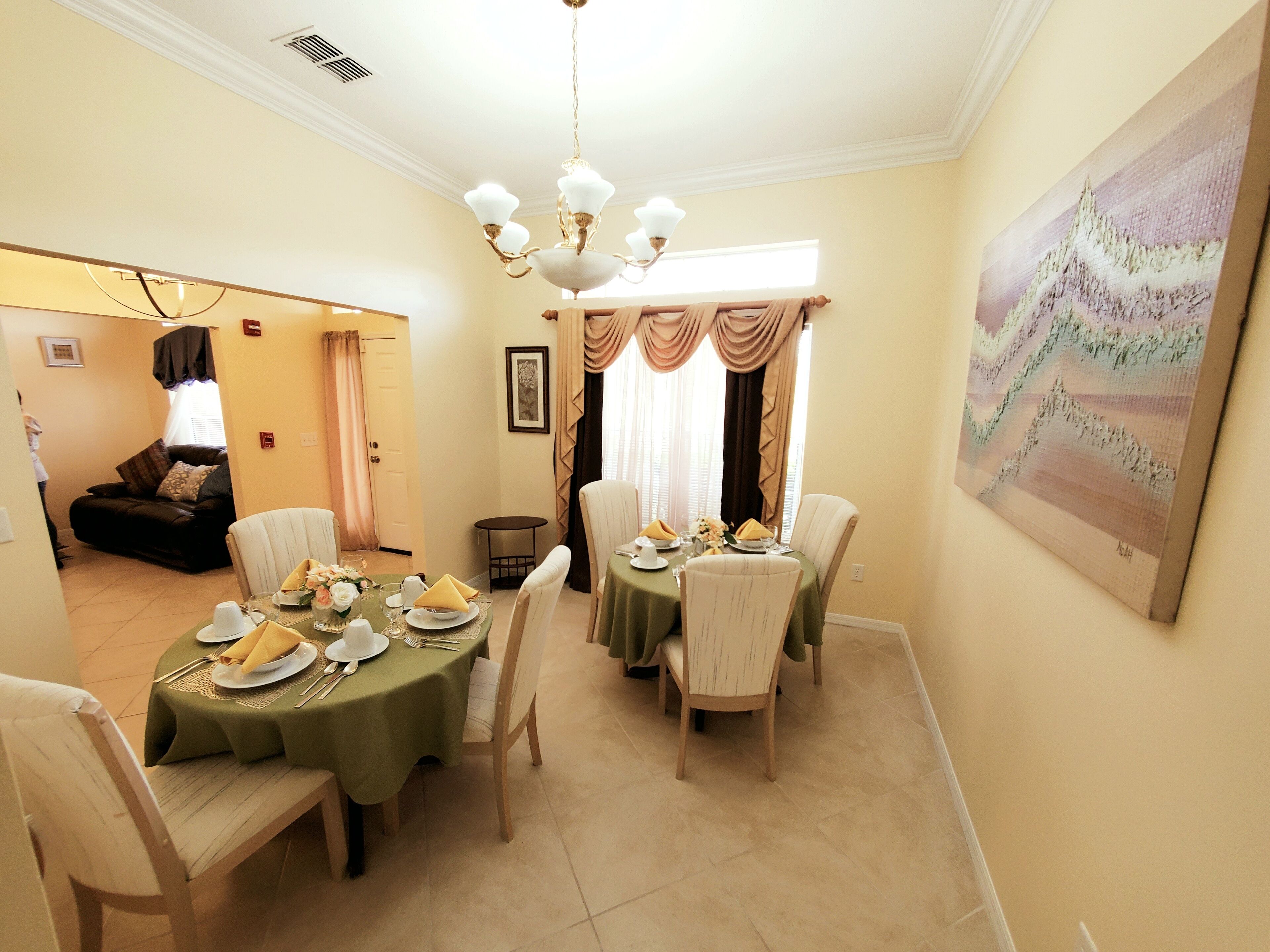 A dining area with a table set for four, chairs, a chandelier, and a painting on the wall.
