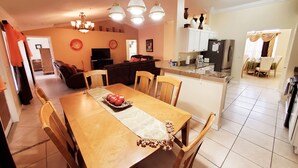 A dining area with a wooden table, chairs, and a decorative centerpiece. There is a kitchen area with cabinets and a refrigerator in the background. - SERENITY Vacation Home • Heated Pool • Sleeps 12 (Port St. Lucie)
