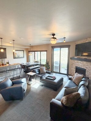 A modern living room with a sectional sofa, a dining area with a table and chairs, a kitchen with bar seating, and a fireplace.