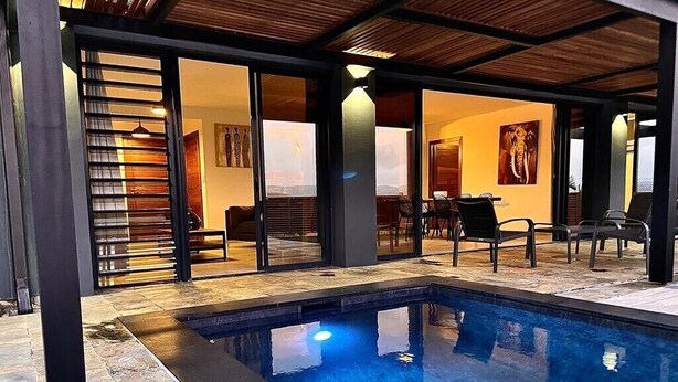 Zona de piscina exterior moderna amb un solàrium de fusta, un porxo amb vidrieres i una zona de menjador amb cadires i taules.
