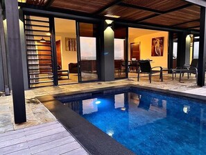 A modern outdoor pool area with a wooden deck, a glass-enclosed patio, and a dining area with chairs and tables. - Luxury Villa Passion Cannelle, private pool, sea and Ilets views (Les Trois-Îlets)