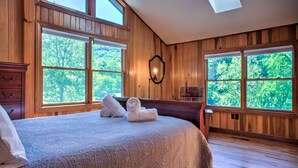 A bedroom with a large bed, wooden walls, and multiple windows offering a view of trees.