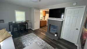 A living room with a fireplace, a television mounted above it, and a dining area with a table and chairs. - COZY COTTAGE NEAR CEDAR POINT/ KALAHARI /SUMMIT RACEWAY (Norwalk)