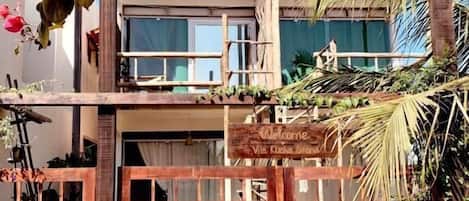 Una puerta de madera con un letrero que dice 'Welcome', un edificio moderno con ventanales grandes y plantas tropicales.