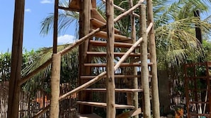 Una escalera de madera al aire libre que sube hacia un espacio cubierto, rodeado de palmeras y follaje.