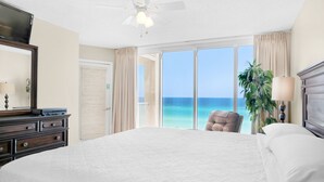 A hotel room with a large bed, a ceiling fan, a view of the ocean, a chair, and a plant.