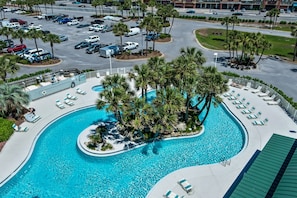 A large swimming pool surrounded by palm trees and lounge chairs, with a parking lot in the background.