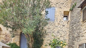 A courtyard with stone steps leading to a door, surrounded by stone buildings with shutters and a tree with yellow flowers.