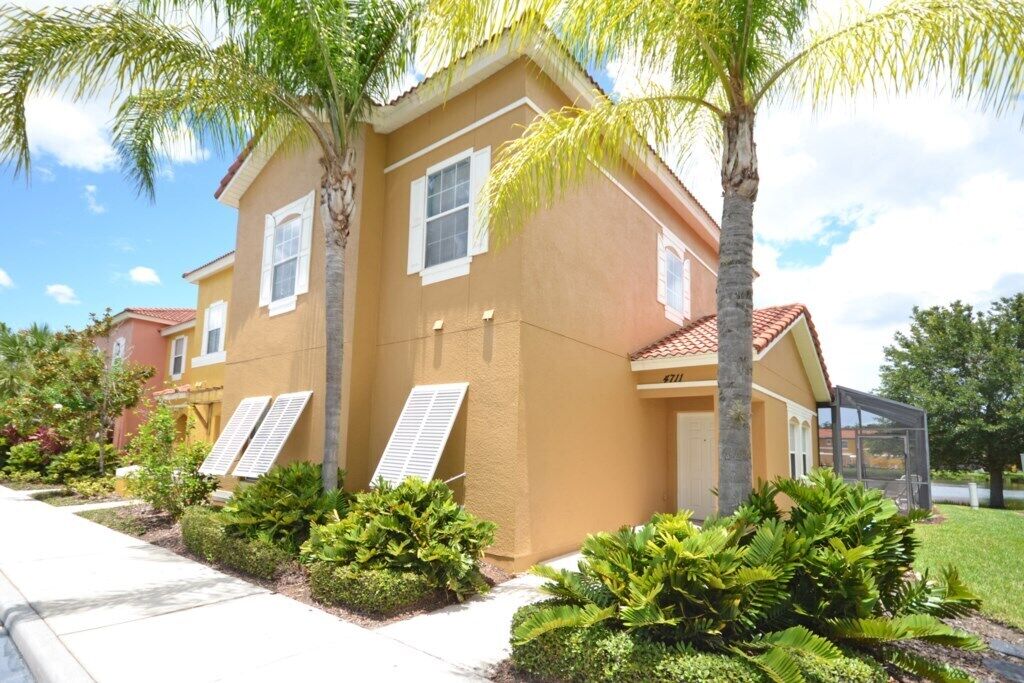 A two-story house with a beige exterior, white shutters, and a red-tiled roof. There are palm trees and well-maintained shrubs in the front yard.