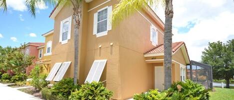 A two-story house with a beige exterior, white shutters, and a red-tiled roof. There are palm trees and well-maintained shrubs in the front yard.
