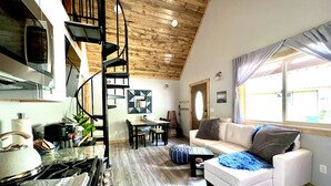Une cuisine moderne avec un escalier en colimaçon, un coin repas avec un canapé blanc et un plaid bleu, et une grande fenêtre avec des rideaux.