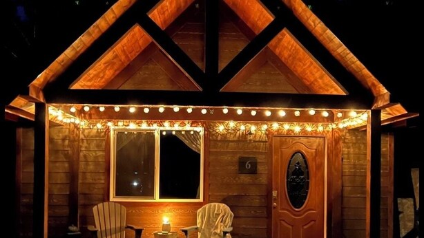 Une cabane en bois avec une véranda, deux chaises, une table et des guirlandes lumineuses.