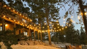 Une cabane en bois décorée de guirlandes lumineuses, entourée d’arbres et bordée d’une zone sablonneuse avec des chaises.