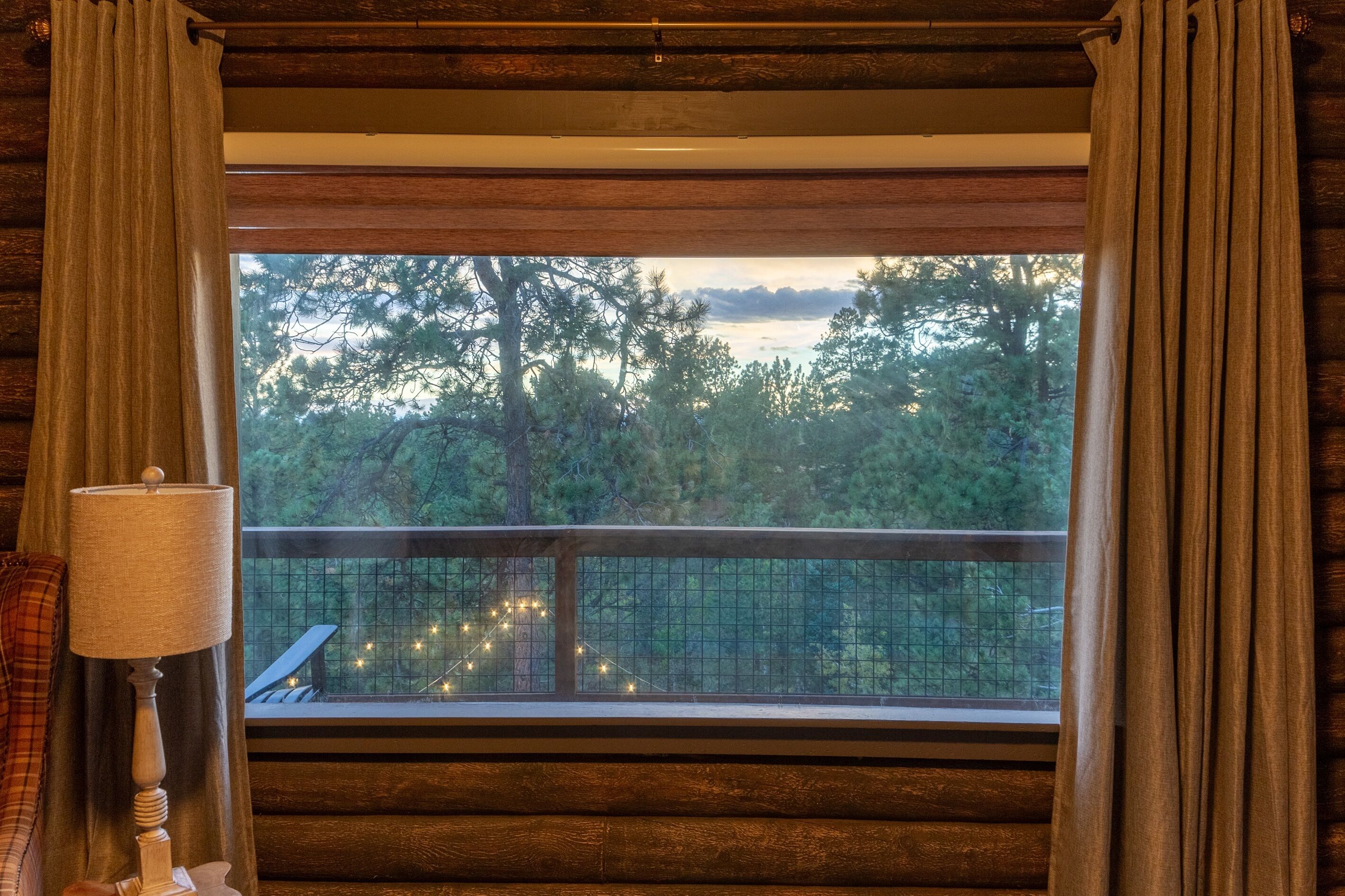 A room with a large window, wooden walls, a lamp, and a view of trees and a balcony with string lights.