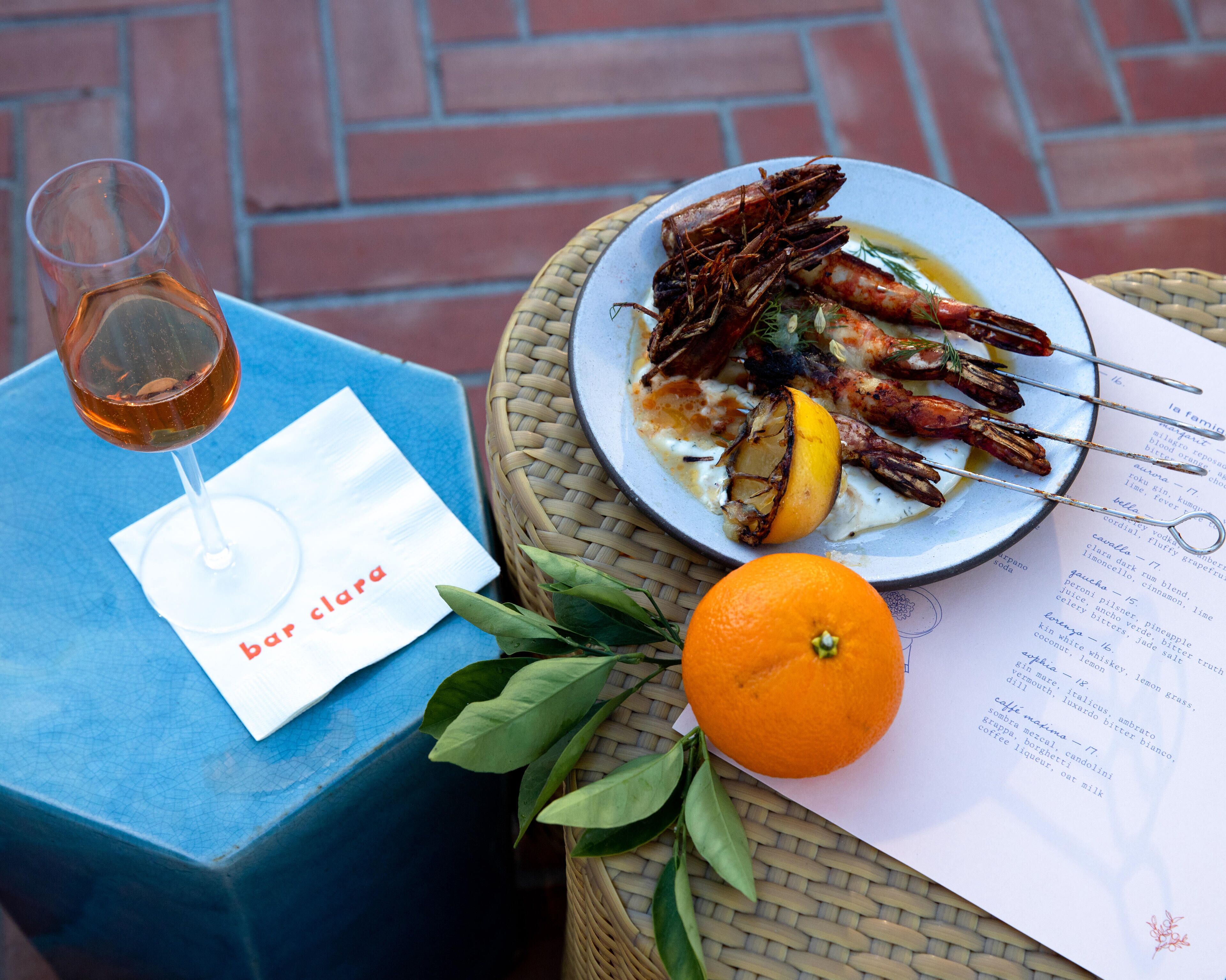 A table with a plate of grilled seafood, a glass of wine, a menu, and an orange.