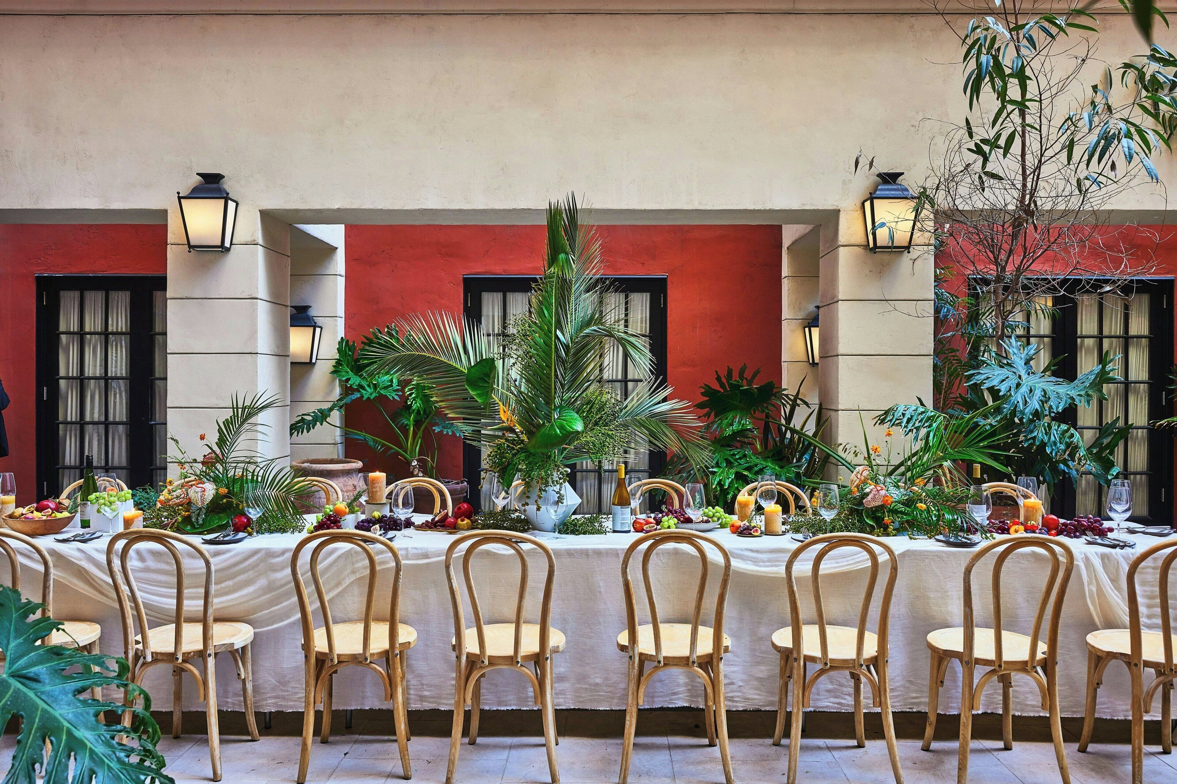 Una larga mesa de comedor dispuesta con sillas, rodeada de plantas tropicales y una variedad de frutas y verduras.