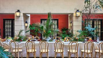 Una larga mesa de comedor dispuesta con sillas, rodeada de plantas tropicales y una variedad de frutas y verduras.