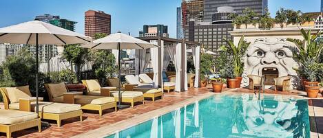 A rooftop pool area with lounge chairs and umbrellas, surrounded by potted plants and a stone sculpture.