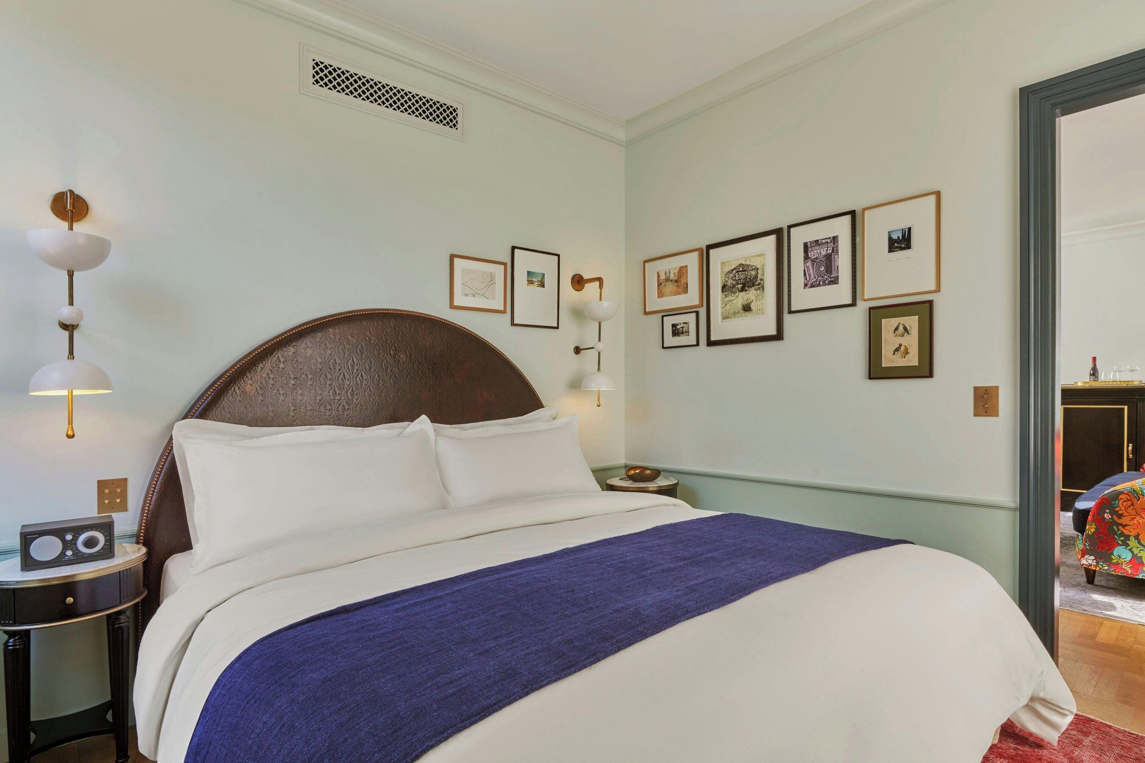 A neatly made bed with a blue blanket, a radio on a bedside table, and framed pictures on the wall.