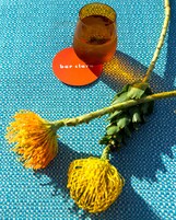 Superficie tessuta blu con un bicchiere di liquido ambrato, un sottobicchiere rosso e un ramo con fiori gialli.