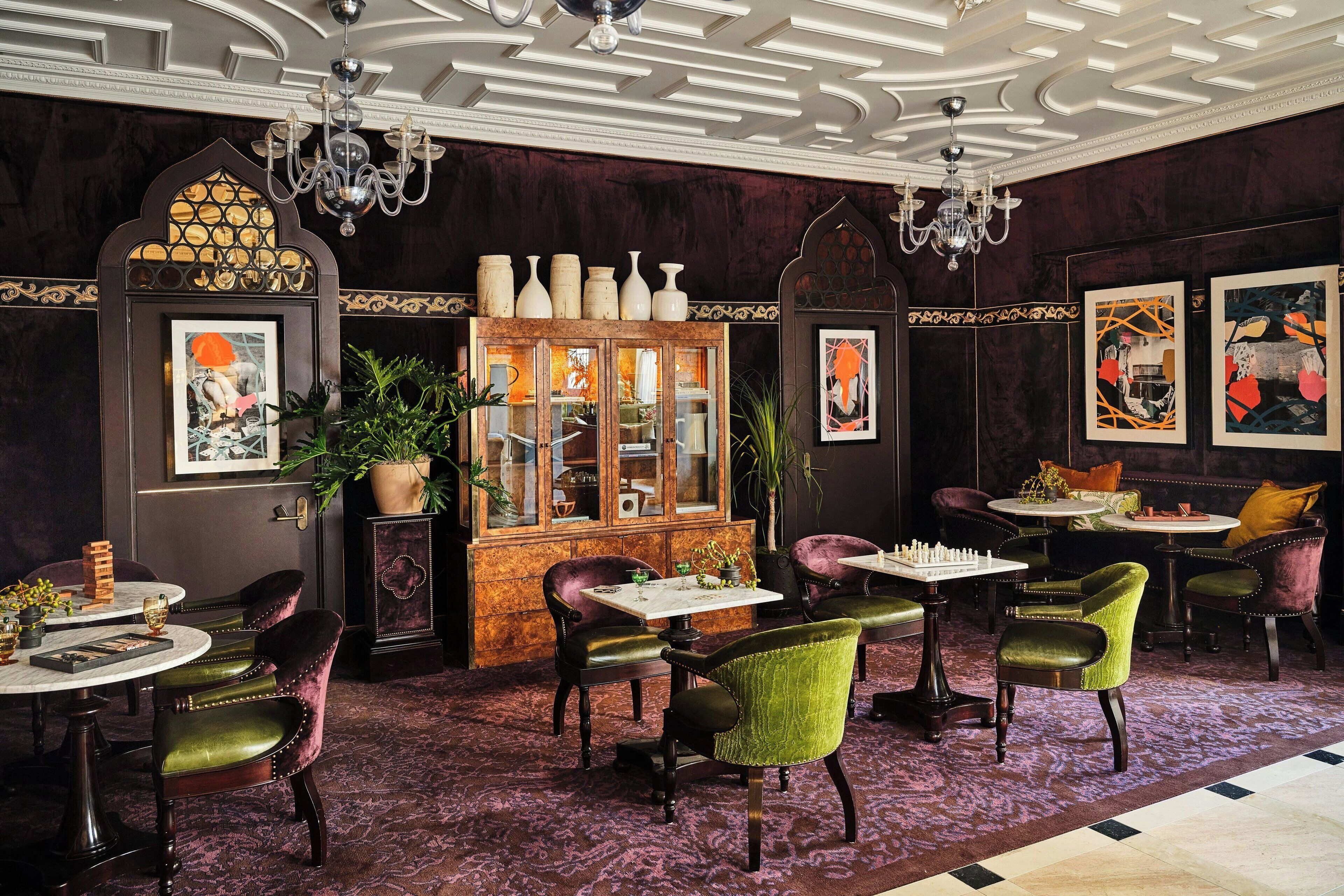 Ein Zimmer mit einem Schachbrett, grünen und violetten Polsterstühlen, einer Vitrine mit Glastüren und einer Chandelüster.